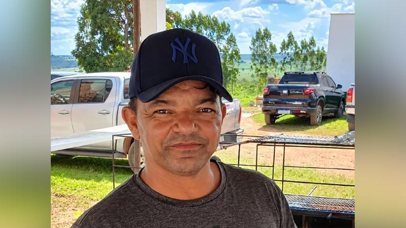 Homem de meia-idade usando boné escuro da marca New York Yankees em uma área externa com picapes ao fundo.