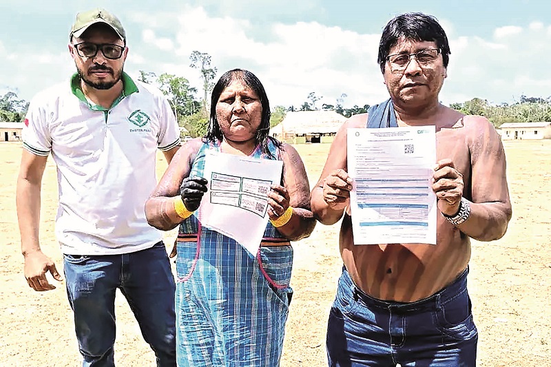 Três pessoas em pé em uma área aberta, com um homem de camisa branca à esquerda e um casal indígena ao lado segurando documentos.
