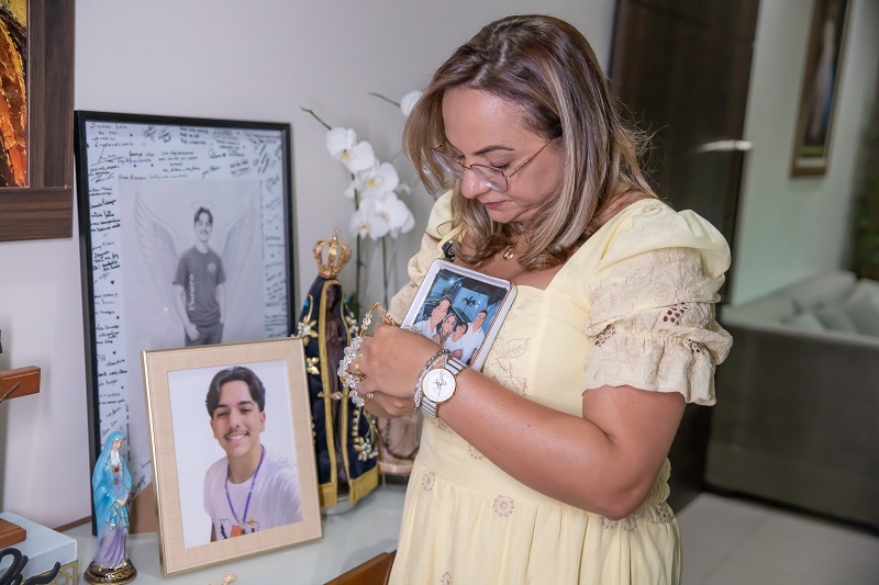 Mulher de vestido amarelo abraça porta-retrato com foto de família em um ambiente com objetos religiosos e porta-retratos de um jovem.